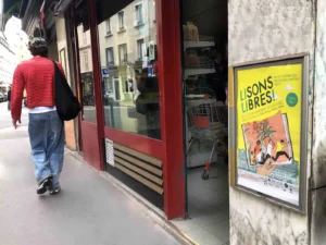 Affiche « Lisons Libres ! » dans un cadre store front sur la devanture d'un commerce parisien, avec un piéton passant devant