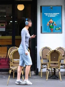 Cadre d'affichage 60 × 80 cm fixé sur la façade d'une brasserie parisienne, affichant une publicité pour le Parc Astérix, devant un piéton qui passe