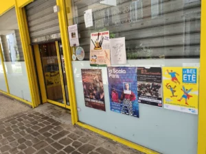 Affiches de concerts et d'événements culturels collées au ruban adhésif sur la vitrine d'un commerce aux encadrements jaunes pour les événements : La Maison Tellier, Lou Trotignon, Le Bel Été d'Est Ensemble.