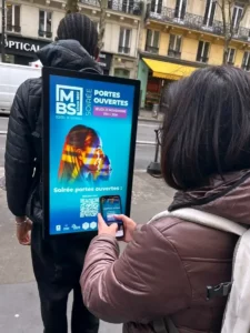 Un marcheur porte un écran LED affichant une publicité et une passante scanne le QR code avec son téléphone. Le LED WALK est un écran publicitaire porté par des piétons pour une présence DOOH mobile.